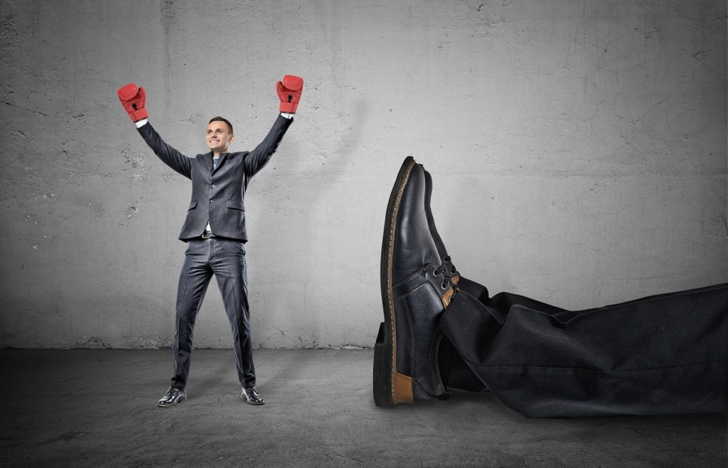 A happy businessman with boxing gloves on arms raised in victory stands near a giant male leg fallen down.