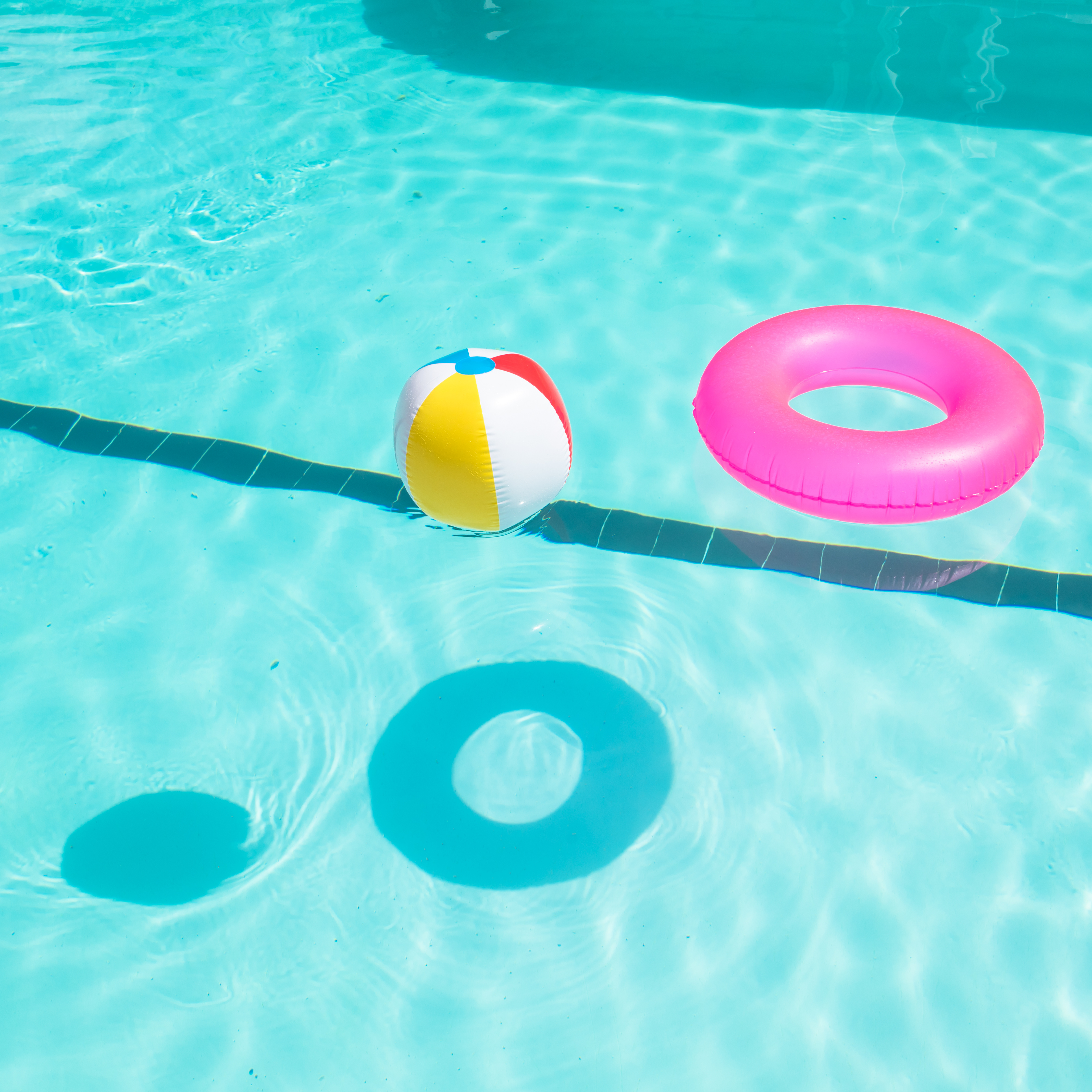 Bright pink float and beach balls in blue swimming pool, floating in refreshing swimming pool with waves reflecting in summer sun. Active vacation background. Lifesaver for kid. Sunny day at the pool