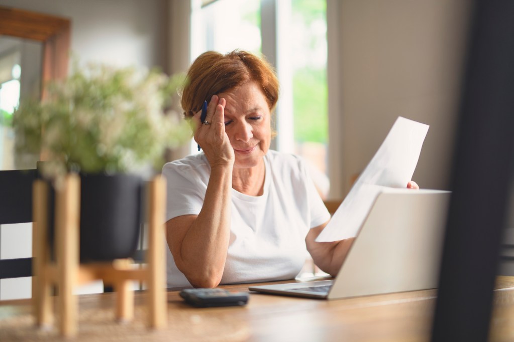 A Sad frustrated senior woman pensioner having depressed look, holding hand on her face, calculating family budget, sitting at kitchen counter with laptop, papers, coffee, calculator and cell phone